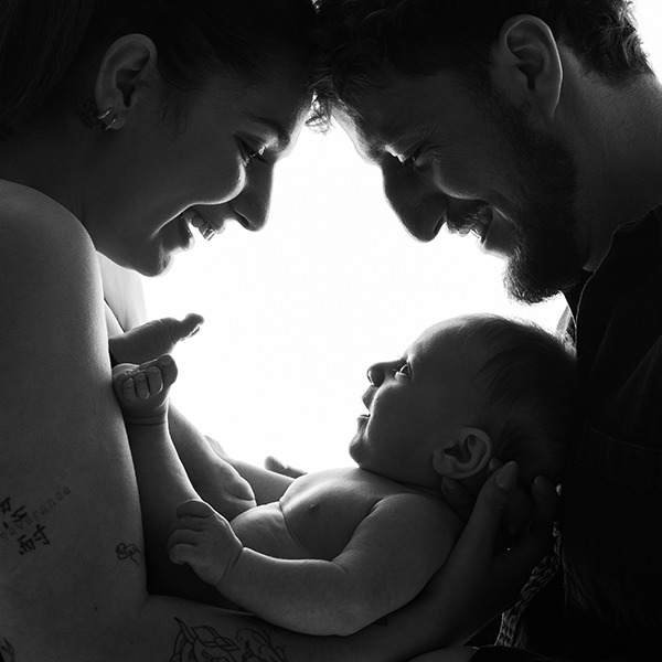 Bébé qui sourit avec tendresse dans les bras de ses parents, il a deux mois. La séance a lieu dans le Studio olivier Leleu à Frontignan